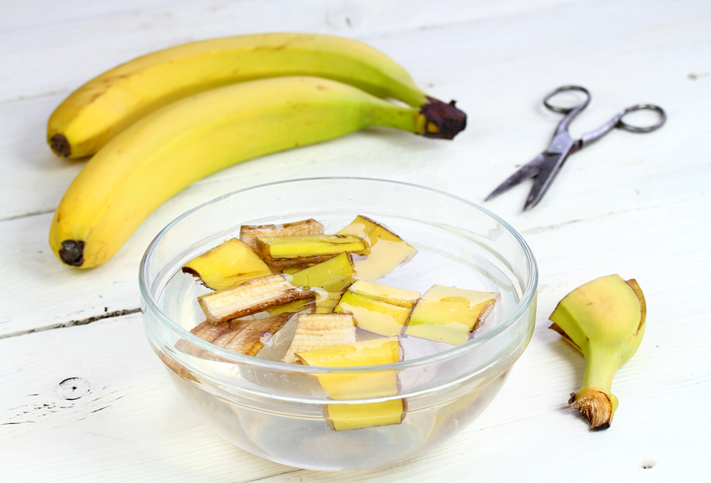 Două banane pe o masă albă alături de un bol transparent cu apă și coji de banane tăiate cu foafeca de lângă bol. 