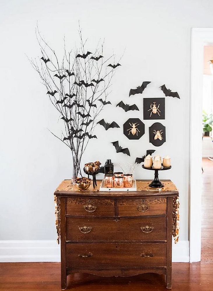 Decorare casă Halloween 2024: o vază transparentă cu crenguțe negre cu lilieci, pe o comodă vintage din lemn, pe un fundal de perete gri deschis, alaături de alte decorațiuni. 